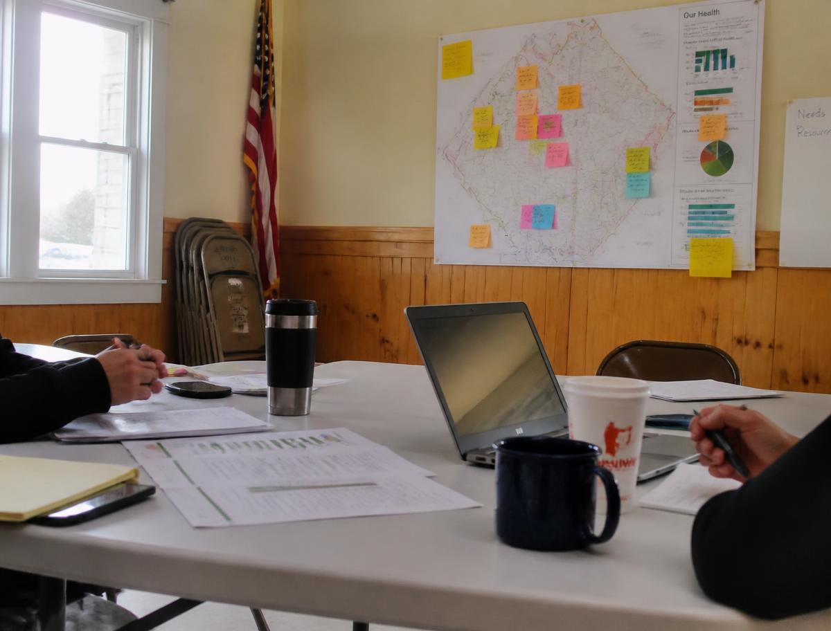 A community center table with printed local health data sheets, coffee cups, and sticky notes on a county wall map during a planning meeting