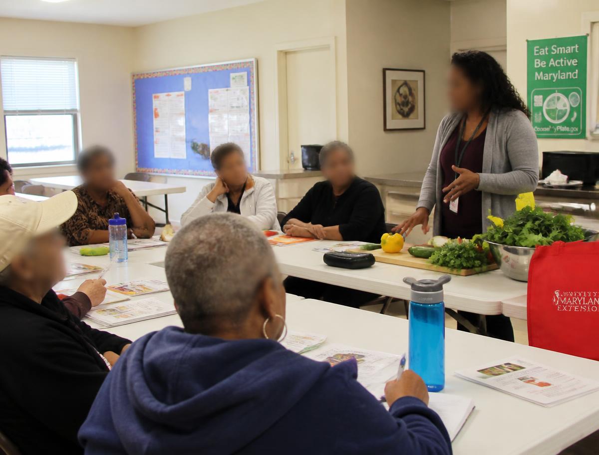 Diverse adults and older adults participate in a community nutrition education class around tables with fresh vegetables, recipe cards, and water bottles